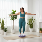 Woman in teal athletic wear standing on a purple balance board in a bright room with plants.